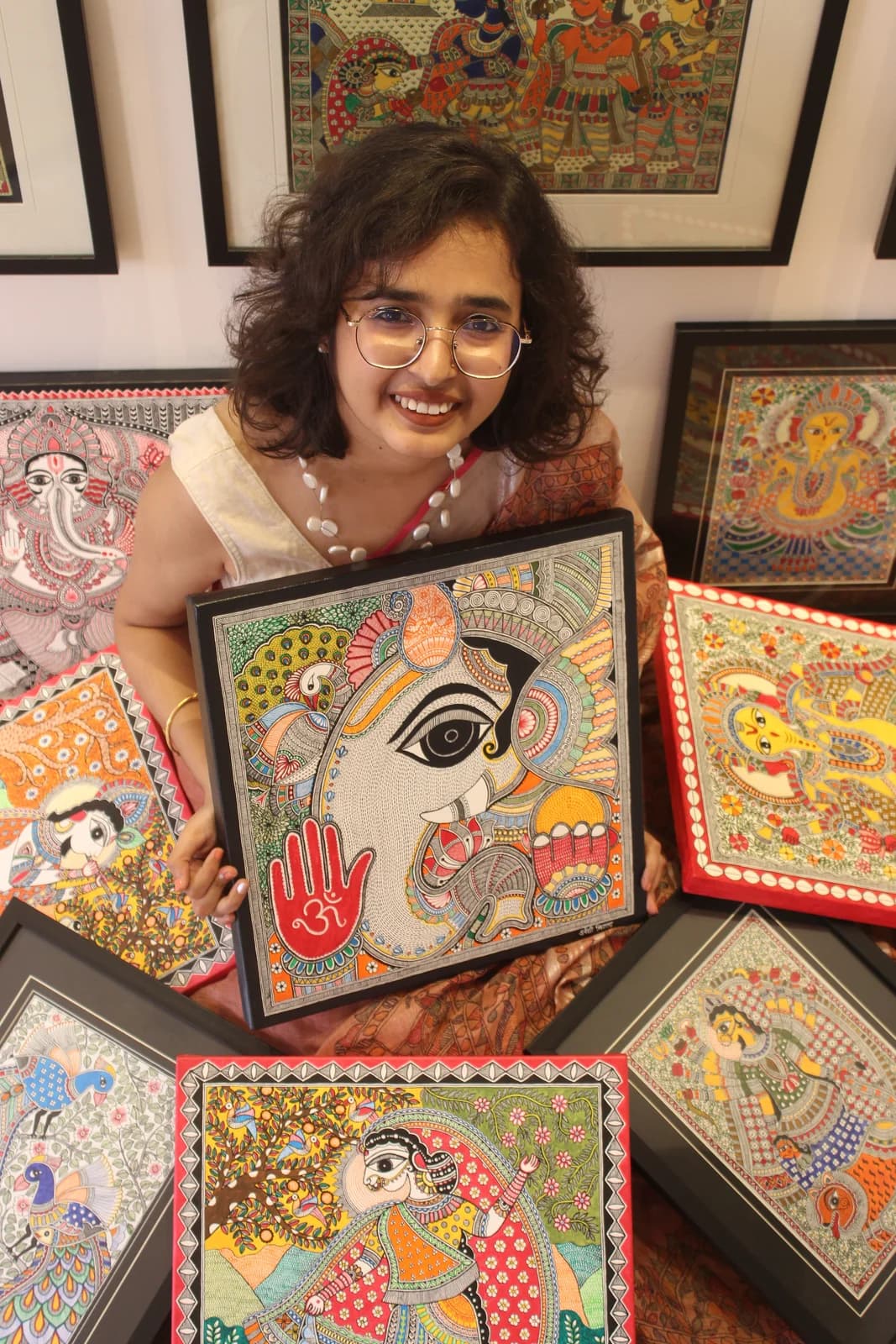 Urwashi Nirala holding a large hand-painted Madhubani artwork of Ganesha, surrounded by more framed paintings in her studio