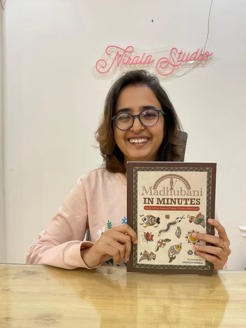 Urwashi Nirala smiling at her Puri studio desk, holding her published book Madhubani in Minutes, with the Nirala Studio neon sign on the wall behind her